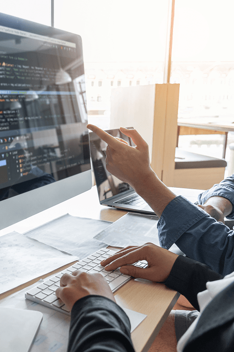 primer plano de dos personas colaborando frente a un monitor que muestra c&oacute;digo de programaci&oacute;n; una persona teclea mientras la otra se&ntilde;ala la pantalla en una oficina iluminada